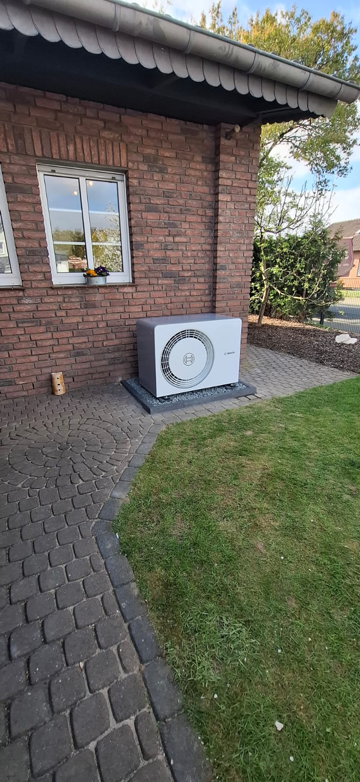 Bosch-Wärmepumpen-Außeneinheit auf Kiesfläche an Backsteinwand unter Fenster, mit sauber verlegten Rohrleitungen.